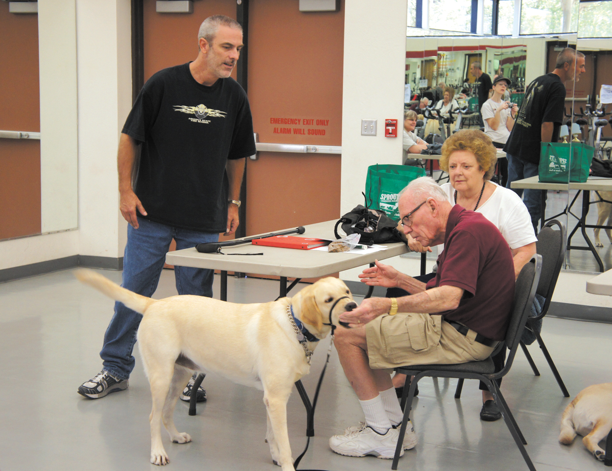 Jake Willison, left, an instructor with Happy Tails Service Dogs, watches as student Robert Westfall commands his 3-year-old yellow Labrador retriever, Molly, to “touch” his hand, while Robert’s wife, Dorothy, looks on. The “touch” command can be used to teach a dog to do everything from turning on a touch lamp to pushing open a door (photo by Teri Carnicelli).