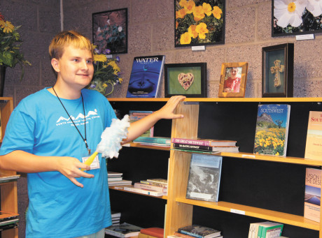 Garrison Mais, 17, dusts the shelves in the library and reading room at the North Mountain Visitor Center, where he’s volunteered for over a year. The top shelf also displays a framed photo of Garrison, with two pieces of his original artwork, also framed, on either side of his picture (photo by Teri Carnicelli).