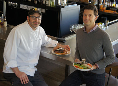 Bernie Kantak, left, chef and partner with The Gladly, and Andrew Fritz, partner and CEO, show off two of the eatery’s most popular items. Kantak is holding The Wagyu Burger, featuring bacon-onion marmalade, Point Reyes blue cheese and arugula on a potato bun, accompanied by fried onion ash fingerlings with a garlic-aioli dipping sauce. Fritz offers up a signature dish first developed at sister eatery, Citizen Public House: The Original Chopped Salad, which has a combination of Israeli couscous, sweet dried corn, smoked salmon, asiago, pepitas, tomatoes, currants and arugula with a basil-ranch dressing (photo by Teri Carnicelli).